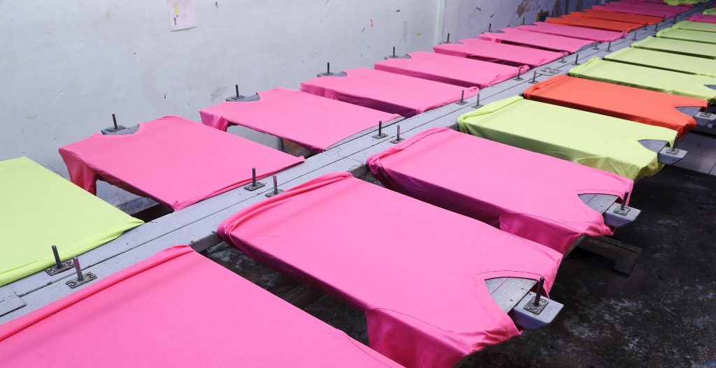 ピンクやライムグリーンなどカラフルなTシャツがプレートに並べられたオリジナルグッズの生産ライン。数量と色・素材が費用に影響する様子が伝わる