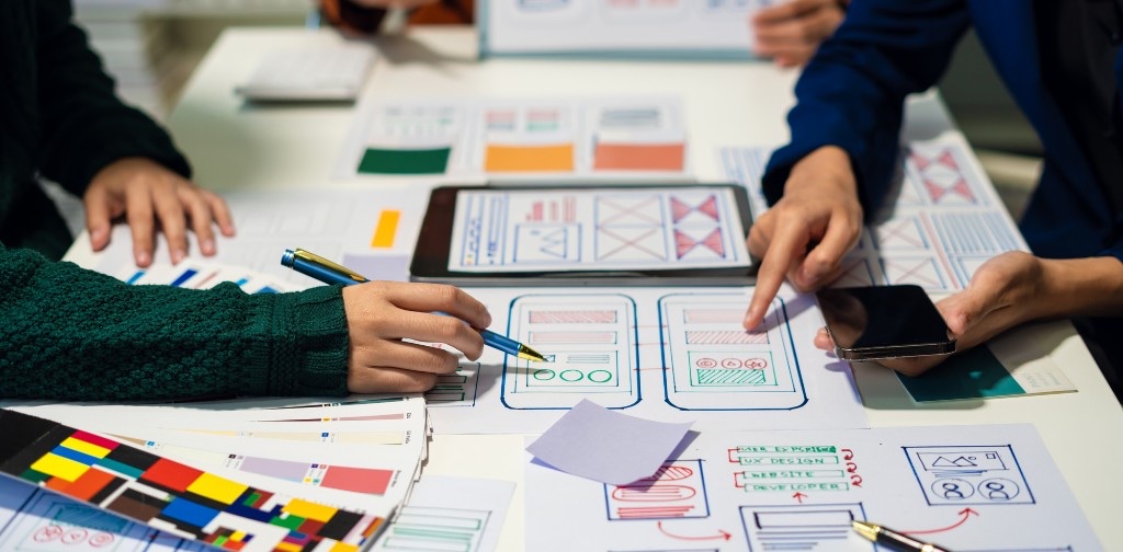 ワイヤーフレームのスケッチとカラーパレットを囲んでデザインを相談する複数人の手元。レイアウトと配色を考える様子