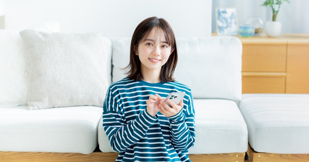 明るいリビングでスマートフォンを両手で操作し、デザインアプリやテンプレを選んでいる女性の写真