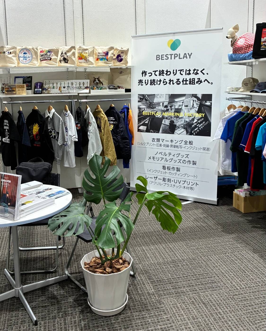 「作って終わりではなく、売り続けられる仕組みへ。」のバナーを掲げたBESTPLAYブースの全景。ウェアやトートバッグ、カタログを展示。